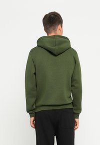 Felpa verde con cappuccio dal taglio rilassato, realizzata in tessuto morbido. Presenta una tasca a marsupio e polsini a costine, mostrata dalla schiena.