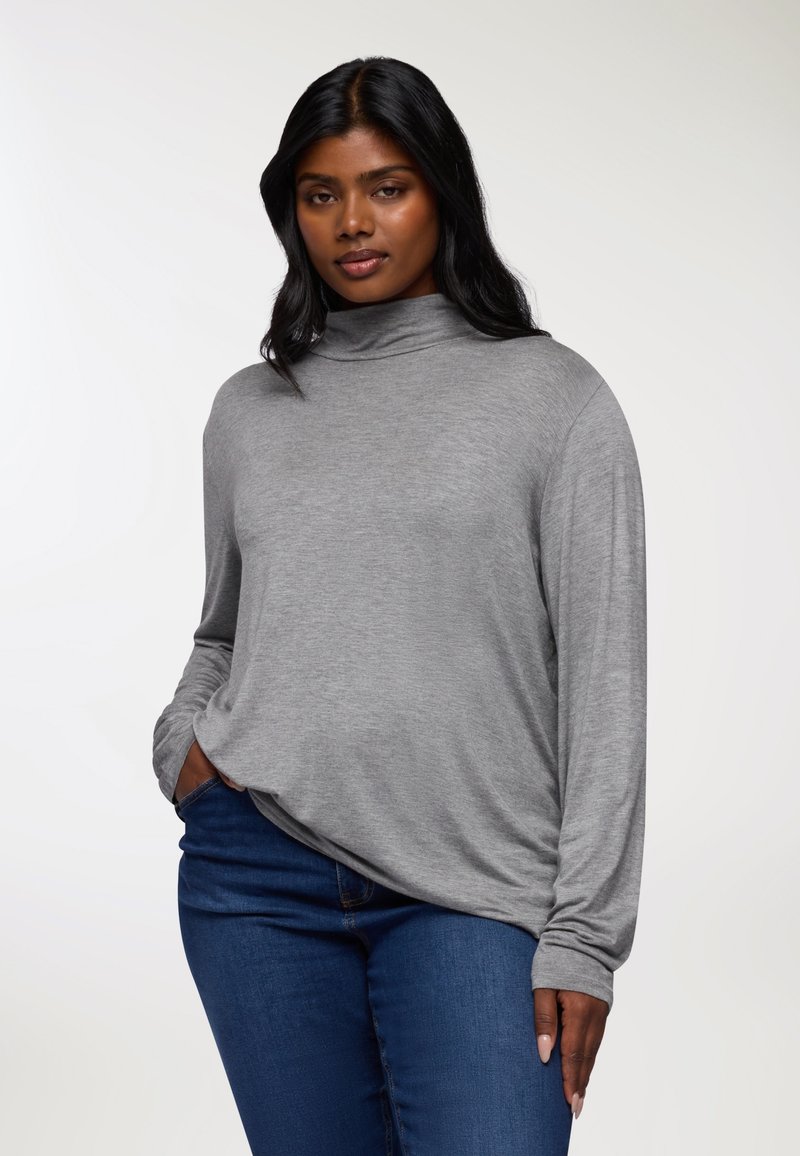 Maglia grigia a maniche lunghe con colletto alto, vestibilità comoda, tessuto morbido e cuciture sulle spalle scese. Abbinata a jeans blu scuro.