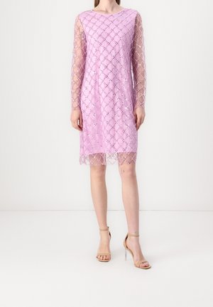 Vrouw draagt een knielange lavendelkleurige jurk met doorschijnende lange mouwen en een diamantpatroon van pailletten, gecombineerd met nude sandalen met hoge hakken.