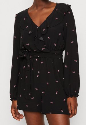 Czarny kombinezon z długim rękawem z różowym nadrukiem paisley, z falbankowym dekoltem w serek, elastyczną talią z wiązanym paskiem, noszony przez osobę o ciemnej karnacji.