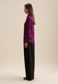 Blusa viola con motivo geometrico, maniche lunghe e taglio sartoriale, abbinata a pantaloni larghi neri, forma slanciata e texture opaca.