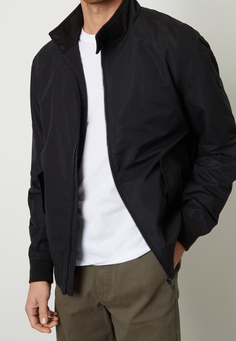 Homme portant une veste zippée noire par-dessus un t-shirt blanc et un pantalon vert olive, une main dans la poche, sur un fond uni.