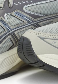 Gros plan de chaussures de sport grises et blanches montrant un tissu en mesh, des surcouches synthétiques, des coutures et des détails de la semelle en caoutchouc texturée.