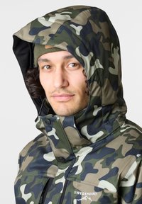 Vihreä ja beige sävyinen camouflage-takki, jossa on korkea kaulus, huppu ja nappikiinnitykset; valmistettu kestävästä, vettä hylkivästä materiaalista.