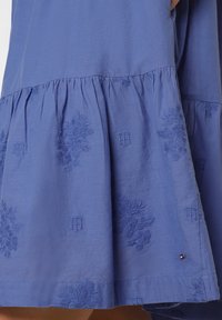 Lila Kleid aus weichem Stoff, mit gestickten Blumenmustern und einem gestuften Design. Enthält dezente Logoelemente nahe dem Saum.