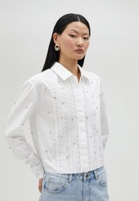 Chemise blanche à boutons avec un devant texturé, comportant des plis verticaux et des embellissements argentés, associée à un jean en denim clair.