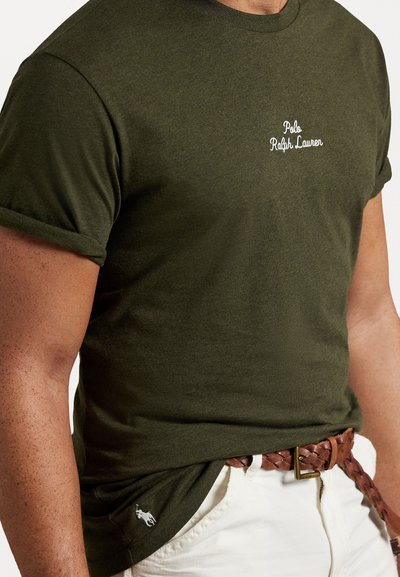 Olīvu zaļš īsām piedurknēm kokvilnas T-krekls ar sarullētām piedurknēm, kura priekšā ir balta izšūta uzraksts "Polo Ralph Lauren" un mazs logo uz apakšmalas.