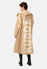 Manteau trench imperméable beige avec un grand texte blanc "KUBOTA" au dos, doté d'une capuche et de longues manches, associé à des bottes noires.