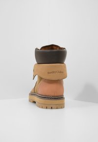 Tan a hnědé kožené kotníkové boty s přehnutým lemem, prošívanými detaily, robustní podrážkou a embosovaným logem na boku.