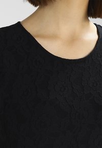 Haut en dentelle noire présentant un motif floral, un col rond et un tissu doux et texturé. Vue en gros plan mettant en valeur le design complexe.