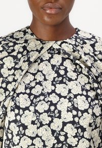 Blommig blus med en mörk marin bakgrund, krämfärgade blommor, hög krage och veckade detaljer. Tillverkad av mjukt tyg med en slät yta.