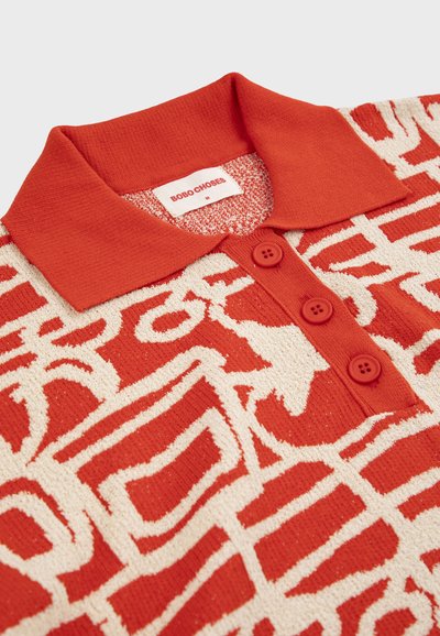 Polo en maille rouge et crème avec un motif géométrique abstrait, trois boutons rouges et une étiquette "BOBO CHOSES" au col.