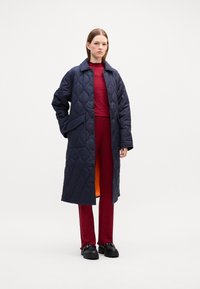 Cappotto trapuntato blu navy con design a colletto, dotato di chiusura a scatto e tasche laterali, indossato su un top rosso testurizzato e pantaloni coordinati.