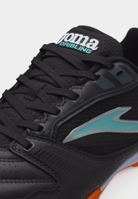 Κοντινή λήψη μαύρου αθλητικού παπουτσιού με λογότυπο σε τιρκουάζ και ασημί, μαύρα κορδόνια και το κείμενο «Joma Dribbling» στη γλώσσα, με πορτοκαλί άκρη στη σόλα.