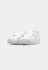 Paire de baskets basses blanches avec lacets à l’avant, semelles épaisses et petit logo doré sur le côté, présentées sur un fond clair uni.