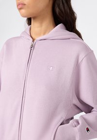 Hoodie di colore viola chiaro realizzato in tessuto morbido, con zip frontale, cappuccio e logo ricamato sul petto e sulla manica.