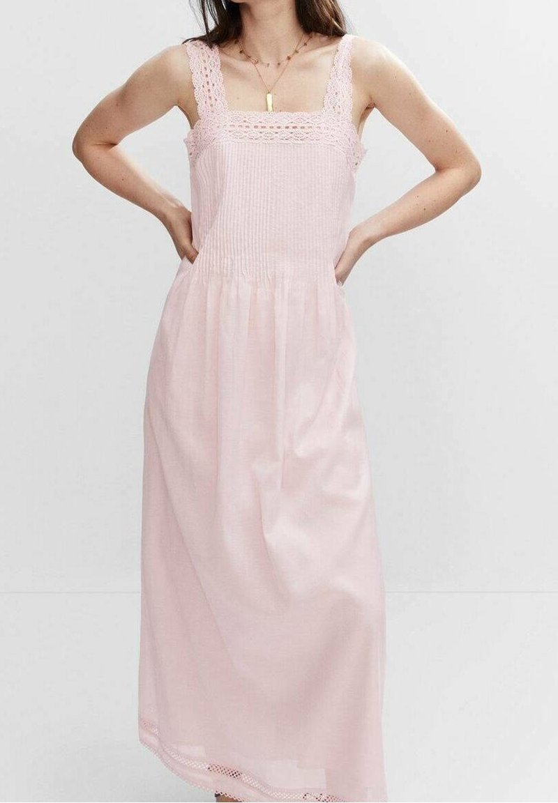 Femme portant une robe midi rose clair sans manches avec des bretelles en dentelle et des détails brodés, debout les mains sur les hanches.