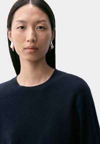 Mörkblå tröja med rund halsringning, gjord av mjukt material. Modellen bär silverfärgade geometriska örhängen, minimalistiska och reflekterande.