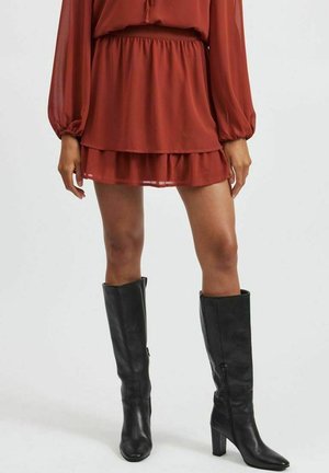 Kvinde iført en rustrød lagdelt miniskørt og sorte læderstøvler til knæet med blokhæle, stående foran en ensfarvet baggrund.