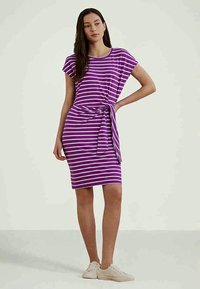 Naine seisab käsi puusal, kandes lillat ja valget triibulist põlvepikkust kleiti ja valgeid tennisesusse ning taust on lihtne.