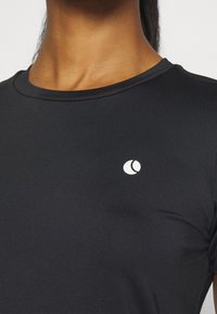 Camiseta deportiva negra con cuello redondo y mangas cortas. Presenta un pequeño logotipo blanco en la parte izquierda del pecho. Textura suave y lisa.