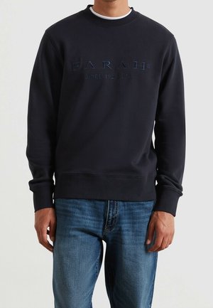 Mand iført en sort sweatshirt med "FARAH SINCE 1920 USA" broderet på fronten, kombineret med blå jeans, mod en ensfarvet baggrund.
