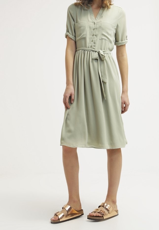 Femme portant une robe boutonnée vert clair avec manches retroussées et ceinture à la taille, associée à des sandales métalliques dorées à boucle.