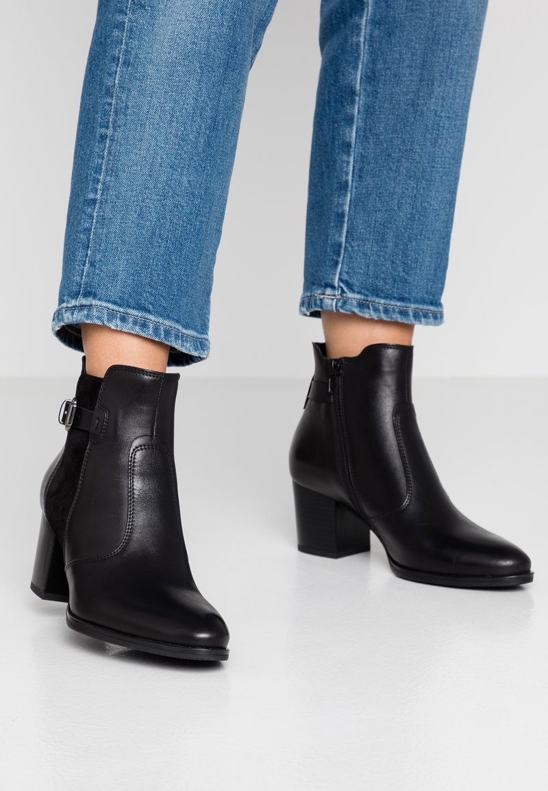 Personne portant un jean bleu et des bottines en cuir noir avec talons carrés et fermetures éclair latérales, debout sur un sol blanc.