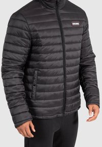 Veste matelassée noire isolée avec un design matelassé horizontal, fermeture éclair à l'avant et poches latérales. Présente une écusson logo sur la poitrine.