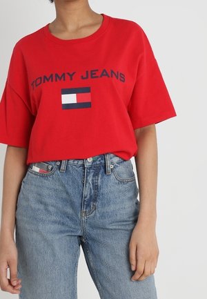 Persoon in een rode "Tommy Jeans" t-shirt met logo en hoge taille blauwe spijkerbroek, staand tegen een effen achtergrond.