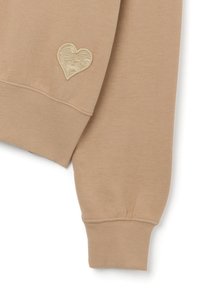 Manica di felpa beige con polsino a coste e un design ricamato a forma di cuore testurizzato in un colore a contrasto. Materiale morbido in misto cotone.