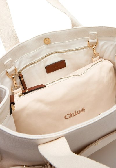Borsa a tracolla in canvas color crema con interno beige, dotata di una tasca con zip, dettagli in pelle marrone e un logo dorato impresso sulla fodera.