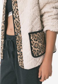 Sølvcreme jakke med leopardmønstret lomme, trykknapper og sorte detaljer; parret med mørkegrå sweatpants.