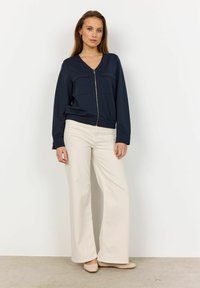 Navy zip-up jack met een V-hals ontwerp, twee borstzakken en een elastische tailleband, gecombineerd met crèmekleurige, wijde broek en beige flats.