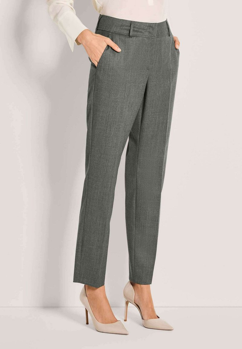 Vrouw in grijze op maat gemaakte broek met zijzakken, gecombineerd met beigekleurige hoge hakken met spitse neuzen, staande tegen een effen achtergrond.