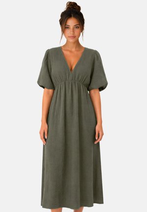 Femme avec les cheveux en chignon porte une robe ample vert olive avec un décolleté en V profond et des manches courtes bouffantes, debout devant un fond uni.