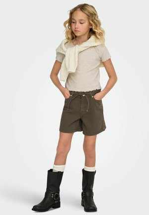 Shorts marron avec une taille élastique, haut gris clair texturé et pull crème drapé sur les épaules. Bottes noires montantes avec boucles.
