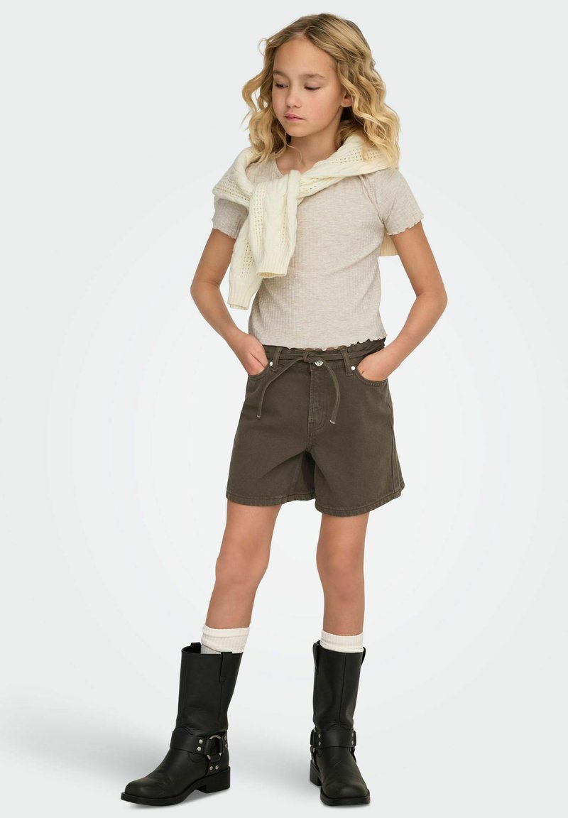Shorts marron avec une taille élastique, haut gris clair texturé et pull crème drapé sur les épaules. Bottes noires montantes avec boucles.