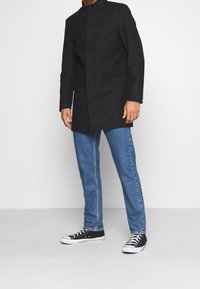 Černý kabát s dlouhými rukávy a vysokým límcem, s bočními kapsami a rovný střihem. Kombinován s modrými džíny a černými teniskami.