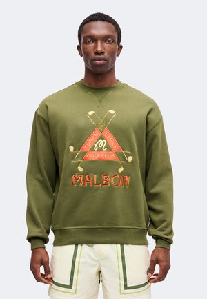 Mann trägt olivgrünes Sweatshirt mit gekreuzten Golfschlägern und "Malbon Cuban Links Golf Club"-Logo, kombiniert mit weißen Shorts mit grünen Streifen.