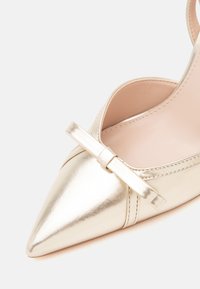 Chaussure plate pointue en métal doré avec une fine bride et un petit détail de nœud à l'avant.