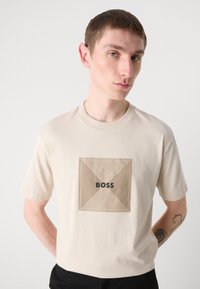 Beige puuvillane T-särk ruudukujulise, soonikuga beeži plaastriga, millel on must kiri "BOSS". Klassikaline ümmargune kaelus ja lühikesed varrukad.