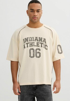 Hombre vistiendo camiseta beige de gran tamaño con el texto "INDIANA ATHLETIC 06" y vaqueros negros desgastados, de pie frente a un fondo claro y sencillo.