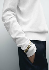 Person som bär en vit långärmad topp med manschetter över svarta byxor, hand i fickan, och bär staplade guld- och silverarmband på handleden.