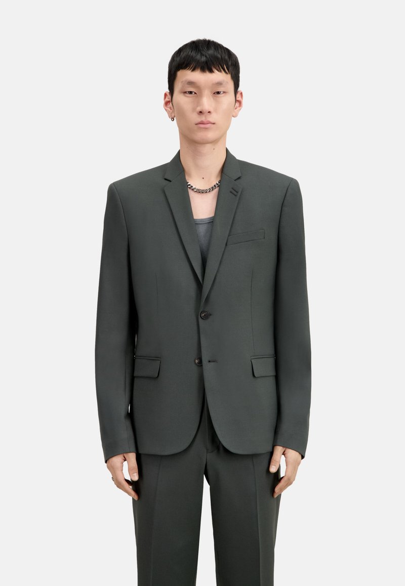 Blazer su misura verde scuro con rever, due bottoni e tasche frontali. Tessuto liscio, vestibilità strutturata, indossato sopra una maglietta grigia.