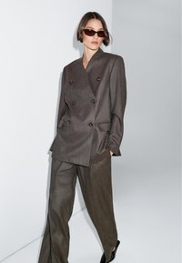 Hnědý dvouřadý blazer s velkými knoflíky, spárovaný s odpovídajícími širokými kalhotami. Hladká textilie, minimalistický design, elegantní silueta.