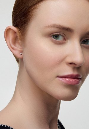 Zbliżenie na kobietę o jasnej karnacji, zielonych oczach, jasnobrązowych włosach zaczesanych do tyłu, noszącą małą diamentową kolczyk w lewym uchu.