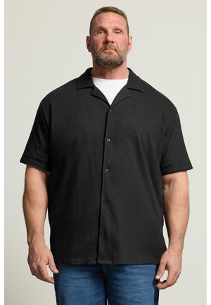 Muskelbepackter Mann mit kurzen Haaren und Bart, der ein schwarzes kurzärmeliges Hemd über einem weißen T-Shirt und blauen Jeans trägt und stillsteht.