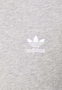 Un tissu en coton gris présente un petit logo Adidas blanc avec trois bandes horizontales au-dessus, positionné au centre du textile.