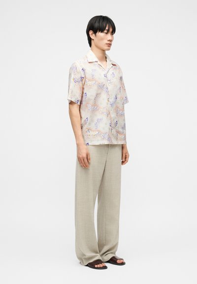 Jeune homme debout portant une chemise florale pastel à manches courtes, un pantalon ample beige et des sandales plates foncées sur un fond uni.